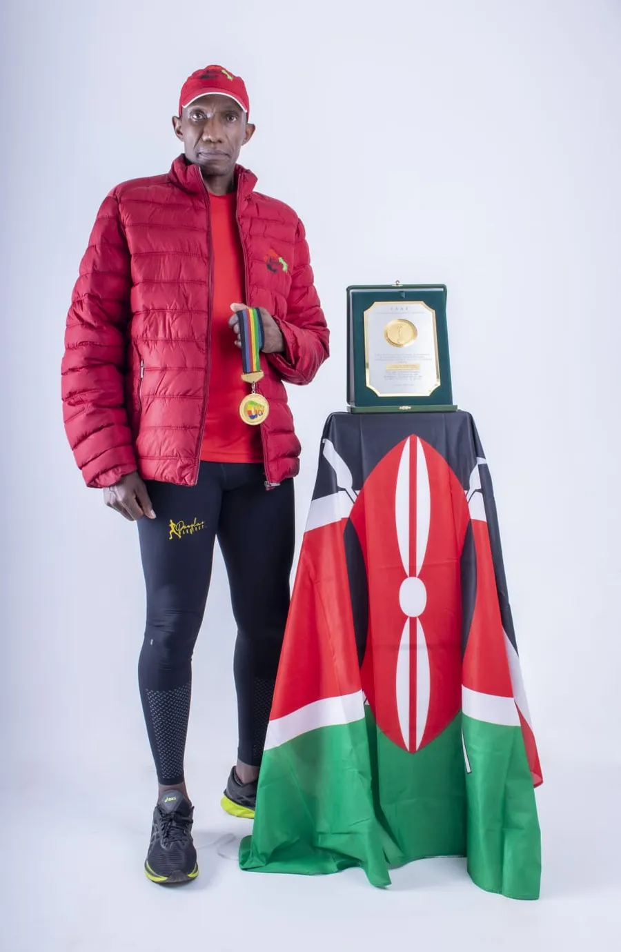 Wakiihuri in a red jacket holding the 1987 gold medal beside the IAAF plaque and a Kenyan flag