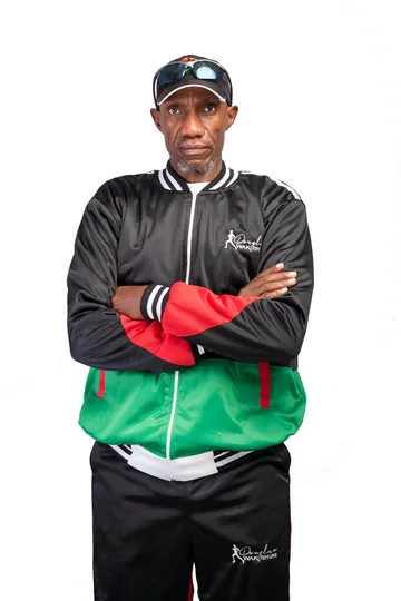 Douglas Wakiihuri in his signature tracksuit, arms crossed, on white